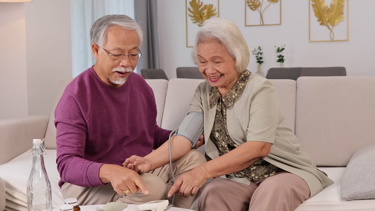 An elderly couple shares a joyful moment while checking blood pressure in a cozy living room setting