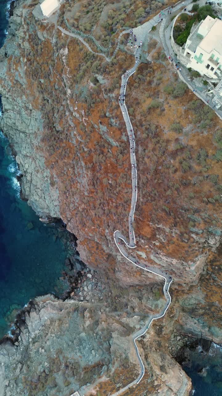 Vertical birdseye drone shot looking straight down at the Church of the Seven Martyrs and the winding stone path on a rocky peninsula in Sifnos island surrounded by the Aegean sea