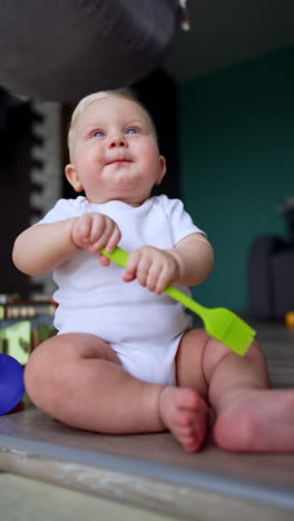 lovely baby in white bodysuit sits on the floor. cute infant plays with kitchen silicone brush. vertical video.