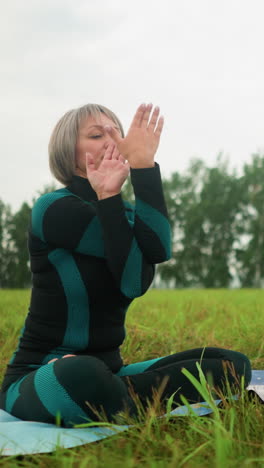 mujer sentada en una alfombra de yoga en un campo brumoso practicando yoga con los ojos cerrados, girando la mano y tocando el pulgar la frente, el telón de fondo incluye un cielo nublado con árboles alineados a lo largo del horizonte
