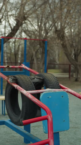 primer plano de una instalación de gimnasio al aire libre con vistas a equipos de ejercicio azules y rojos hechos de neumáticos y barras de metal reutilizados, con árboles estériles y bancos cercanos