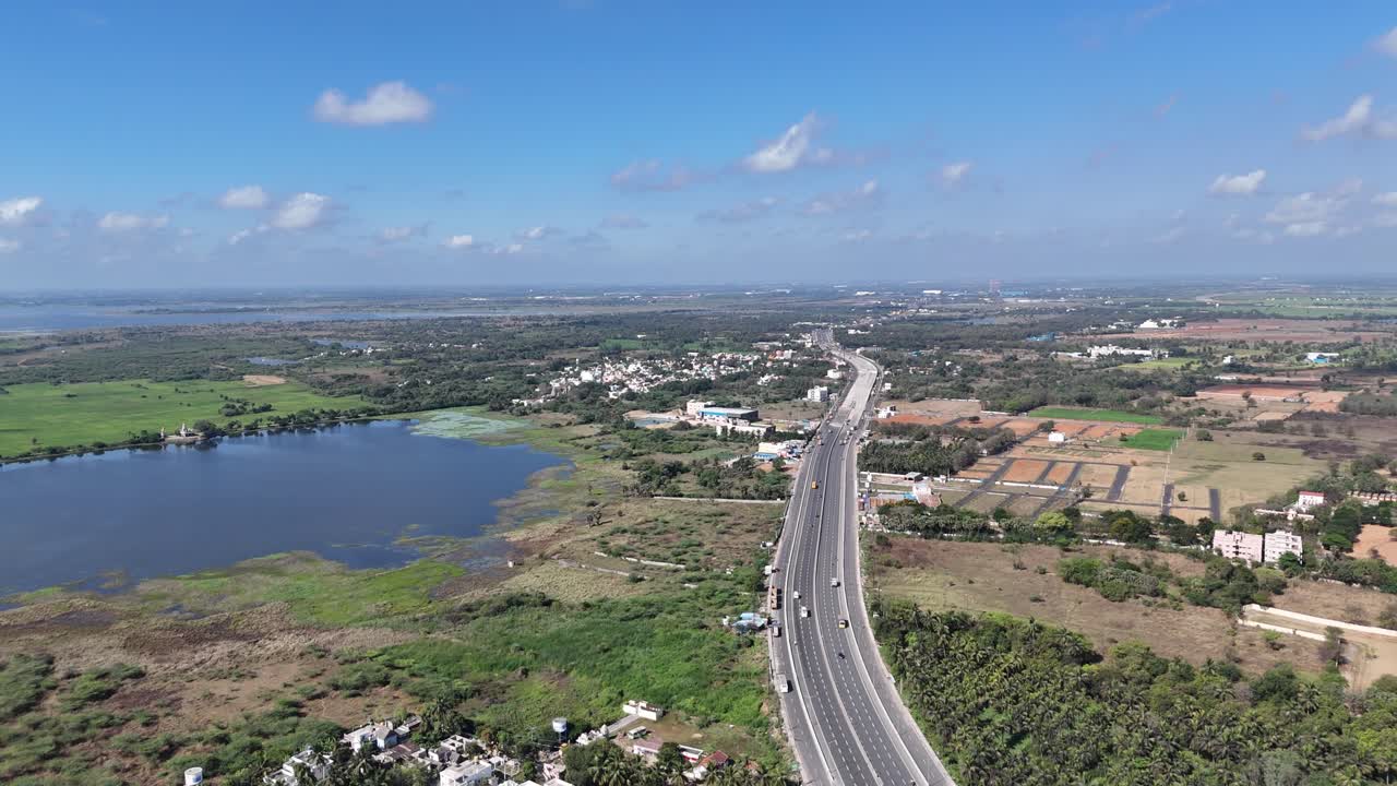 Aerial view: Multi-lane highway construction near Chennai, with earthworks, machinery, and active traffic. It traverses residential areas, fields, undeveloped land, and a large body of water