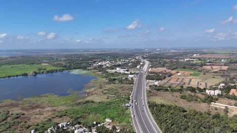 aerial view: multi-lane highway construction near chennai, with earthworks, machinery, and active traffic. it traverses residential areas, fields, undeveloped land, and a large body of water