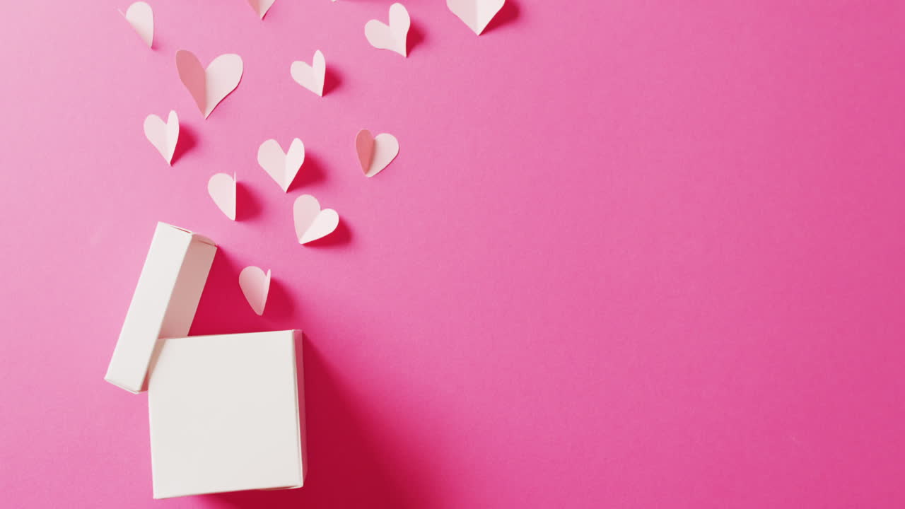 Video of open white gift box and white paper heart shapes, on pink background with copy space
