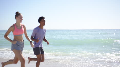 a young caucasian woman and a biracial man, dressed in athletic wear, are jogging along the beach