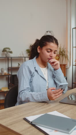 smiling young woman freelancer sitting at table using smartphone texting in living room at home