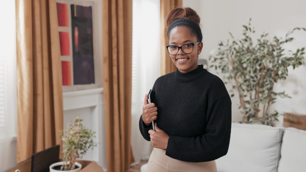 An elegant woman holds a tablet under her arm in hands, looks into the camera with a smile, stands in the middle of new apartment, fresh from moving
