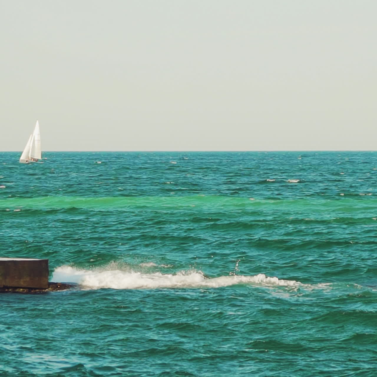 A sailboat, yacht on the horizon in the sea. Splashing waves, ship in the background. Breakwater.