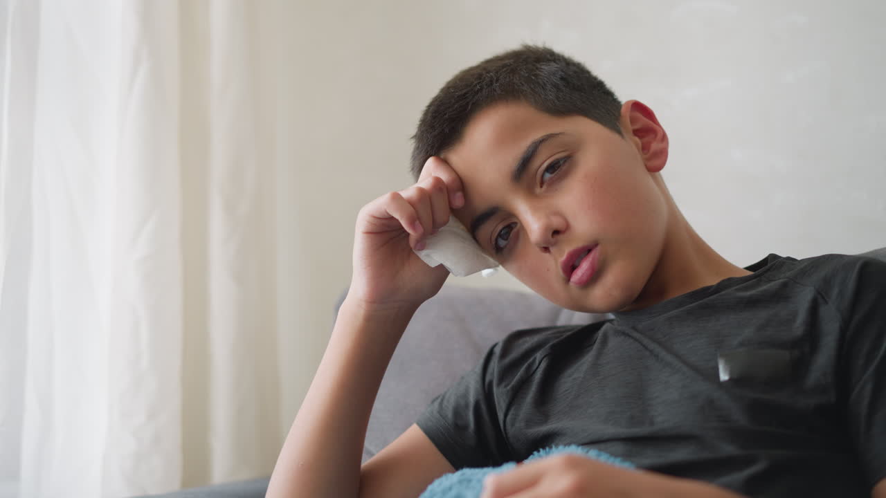 Pale boy looking weak, head resting on hand holding tissue, lying on couch with blanket, looking exhausted and unwell in a home setting with soft natural light filtering through window