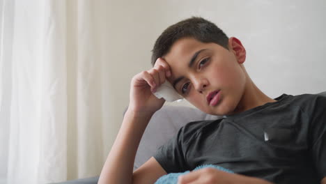 pale boy looking weak, head resting on hand holding tissue, lying on couch with blanket, looking exhausted and unwell in a home setting with soft natural light filtering through window