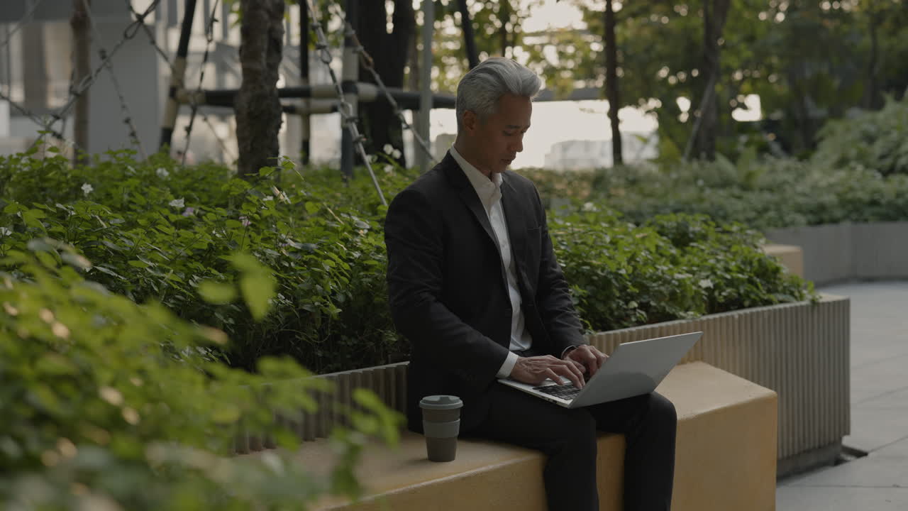 Mature Asian Businessman Working on Laptop Outdoors in Urban Park