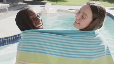 Two-smiling-diverse-teenage-female-friends-covered-by-towel-sitting-by-swimming-pool,-slow-motion