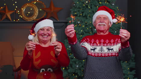 Pareja-De-Ancianos-Con-Suéteres-Festivos-Sosteniendo-Luces-De-Bengala-Disfrutando-De-La-Víspera-De-Navidad-En-La-Habitación-De-Casa