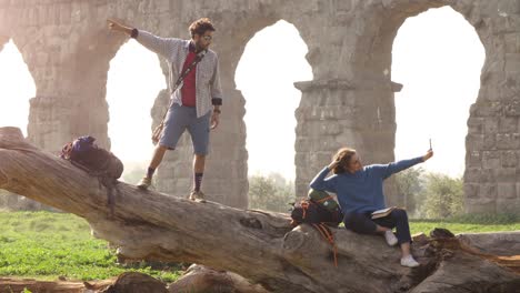 happy young couple backpackers tourists on a log trunk taking selfies photos with smartphone in front of ancient roman aqueduct ruins in romantic parco degli acquedotti park in rome at misty sunrise slow motion tripod