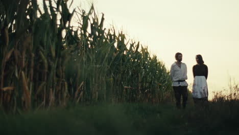 A couple in historical attire standing by a cornfield at sunset