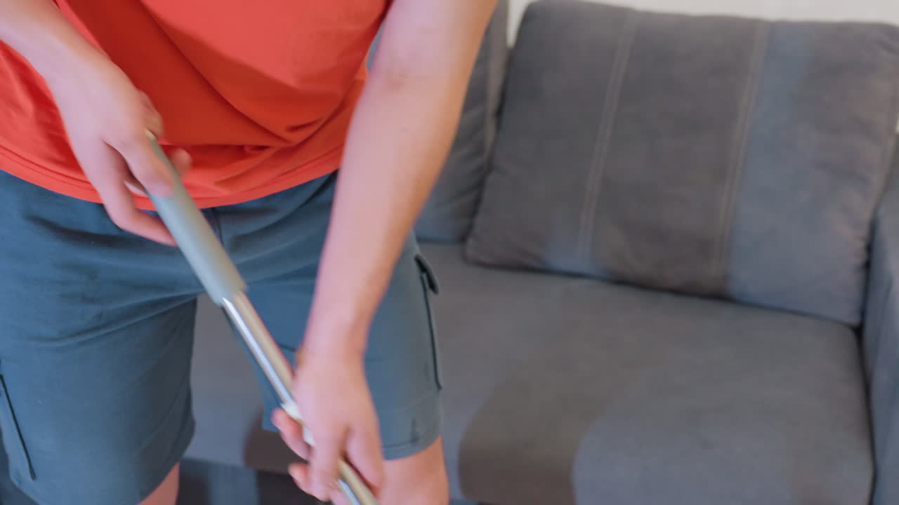 Close up of adult in orange shirt and shorts holding mop handle while cleaning floor near sofa indoors with visible shadow cast on surface showing household maintenance routine