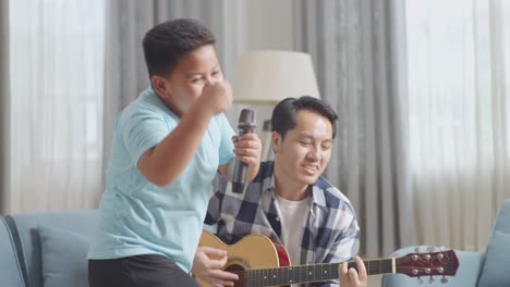 asian father sitting on sofa playing the guitar for his son to dance and sing into microphone at home