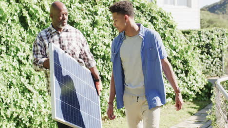 padre afroamericano sosteniendo panel solar hablando con su hijo adulto en un jardín soleado, cámara lenta