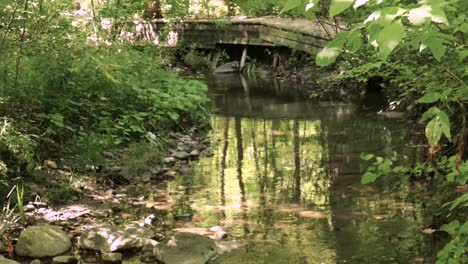 Die-Kamera-Schwenkt-Langsam-Vom-Welligen-Spiegelbild-Eines-Trägen-Baches-Nach-Oben-Zu-Einer-Hölzernen-Fußgängerbrücke