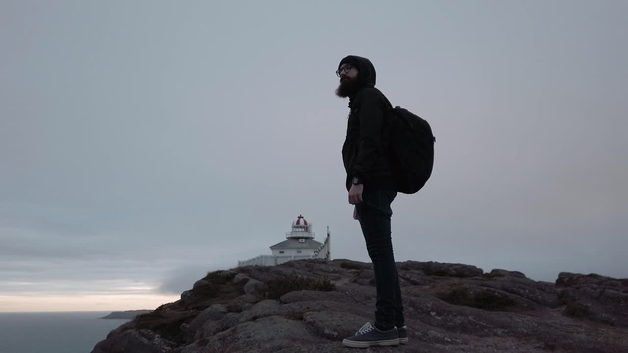 Male hiker with backpack stands on rocky cliff near lighthouse in Canada during cloudy evening, showing solitude, exploration, travel spirit, and scenic coastal environment with calm sea.