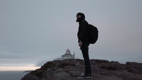 Male hiker with backpack stands on rocky cliff near lighthouse in Canada during cloudy evening, showing solitude, exploration, travel spirit, and scenic coastal environment with calm sea.