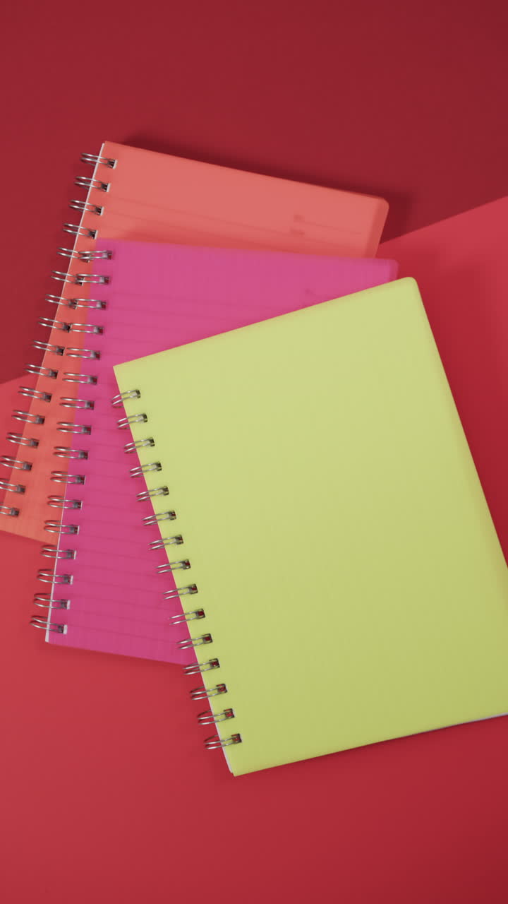 Vertical video of overhead view of colourful notebooks on red background, in slow motion
