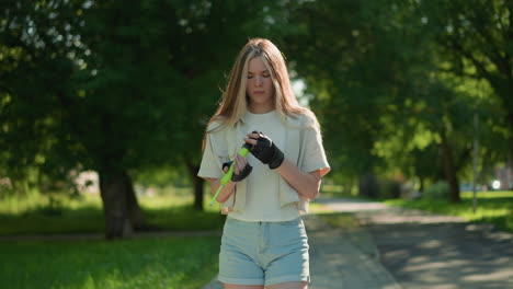 una mujer joven camina al aire libre, examinando una bomba de aire verde mientras la luz del sol se refleja en su cara, está enfocada y pensativa, con un fondo borroso de árboles y un edificio lejano