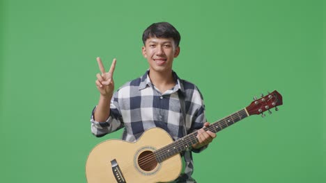 기타를 들고 있는 아시아인 십대가 녹색 스크린 배경에서 평화와 미소를 짓고 있습니다.