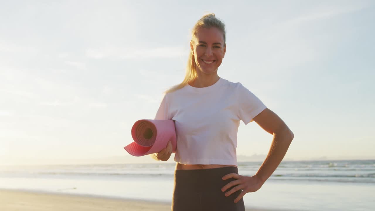 Porträt einer lächelnden kaukasischen Frau, die eine Yogamatte am Strand hält
