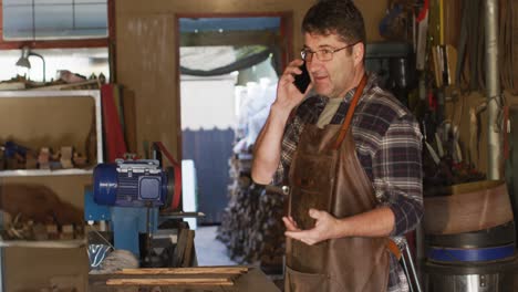 caucasian male knife maker in workshop using smartphone and talking