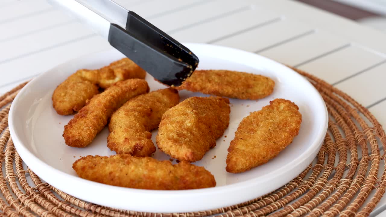 A hand uses tongs to arrange crispy chicken pieces on a white plate. Bright lighting enhances the golden texture