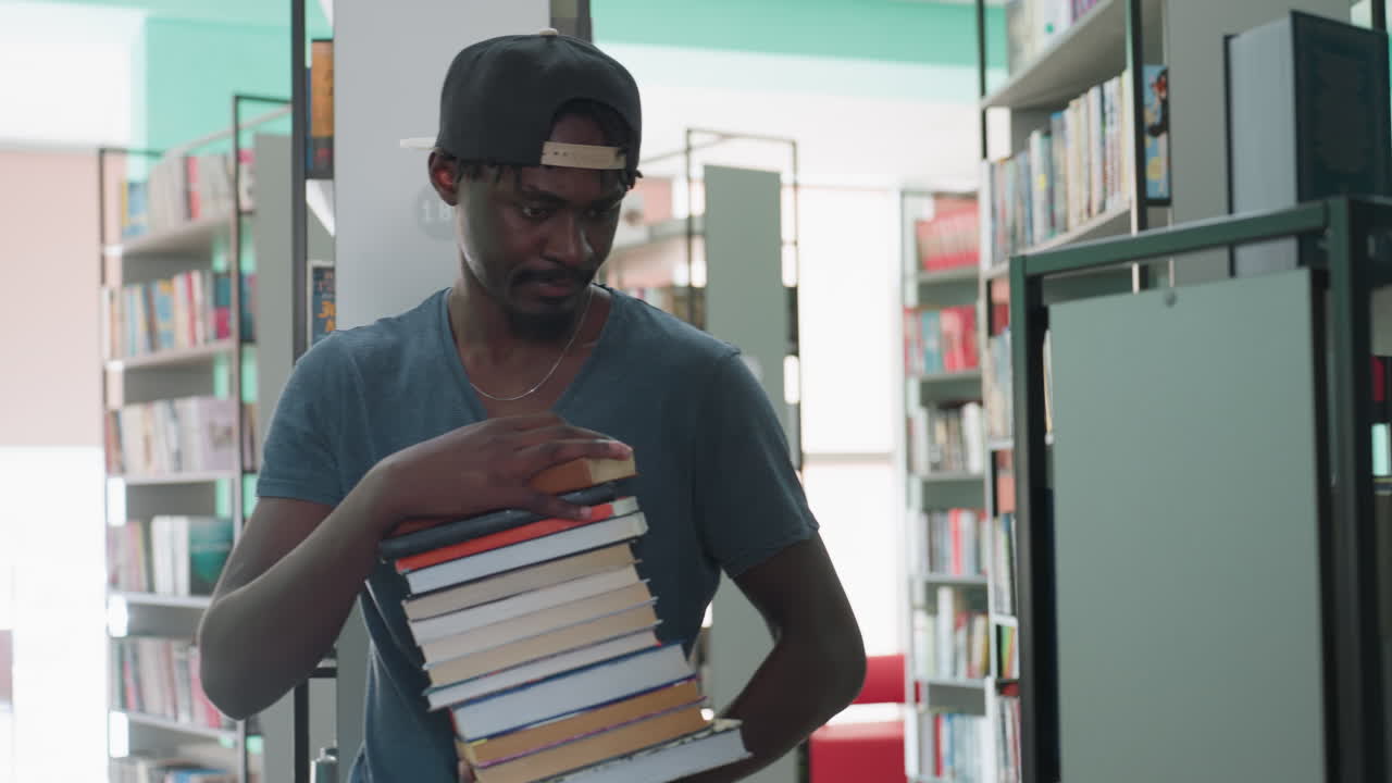 young man in cap enters library aisle carrying tall stack of books, pauses, looks around shelves for spot to place volumes, thoughtful academic vibe with colorful titles