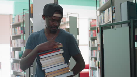 young man in cap enters library aisle carrying tall stack of books, pauses, looks around shelves for spot to place volumes, thoughtful academic vibe with colorful titles