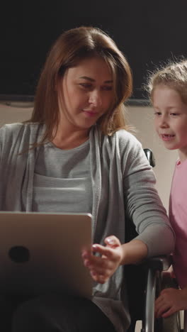 curious children watch freelance mother with leg injury working on laptop at home. woman sits in wheelchair distracting on little daughter and son