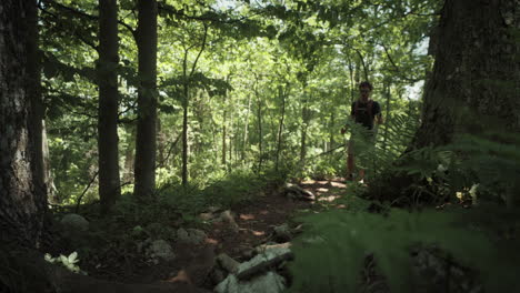 Man-walking-with-a-backpack-and-hiking-poles-up-a-foresty-road,-full-of-rocks,-surrounded-by-bushes