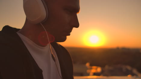 a man in a hoodie sits on the roof and listens to music with headphones typing on a laptop keyboard. run at sunset. freelancer works at sunset with a laptop