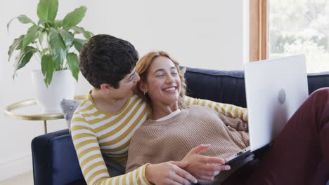 una feliz pareja de lesbianas caucásicas acostadas en el sofá, usando una computadora portátil y riendo en una casa soleada