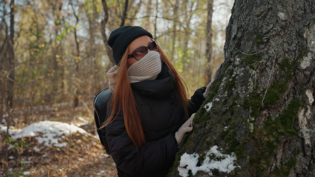 Maskeret kaukasisk kvinde gemmer sig bag træ, spejder ad skovstien som mistænkt for at være en sniger, rødhåret med hue, tørklæde og solbriller, vinterlige sneklatter, mosbegroet bark, spændingsfyldt kold atmosfære