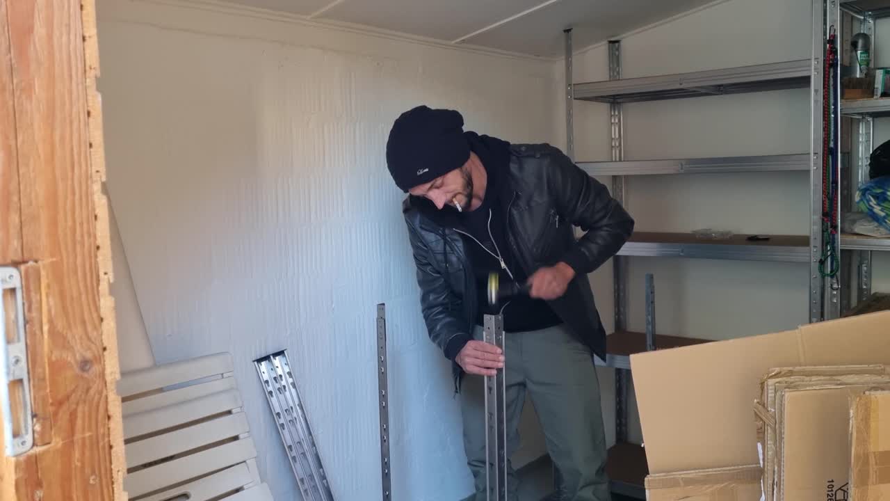 Mann med lue og skinnjakke monterer forsiktig en metallhylle i et kompakt sveitsisk hobbyrom, og nyter et rolig og fokusert gjør-det-selv-øyeblikk hjemme