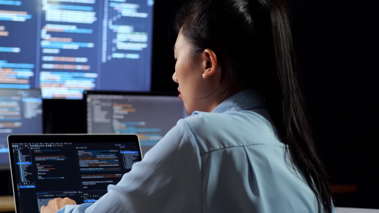 vista cercana de atrás de una programadora asiática apoyándose en la mesa mientras escribe código por una computadora portátil usando múltiples monitores que muestran la base de datos en los escritorios de ventana de terminal en la oficina