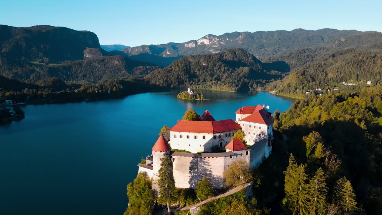 Drone che si allontana in un campo lungo del Castello di Bled in cima alla scogliera all'alba, rivelando il Lago di Bled, la chiesa sull'isola e le Alpi Giulie nella nitida luce del mattino
