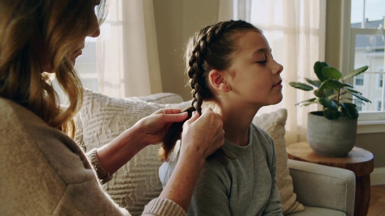 Gathering hair parent finishing Dutch braid on child in living room armchair, knit sweater to tidy