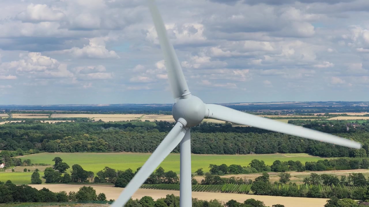 Pale di turbine eoliche ruotano per generare energia pulita sopra i paesaggi di campagna durante un pomeriggio soleggiato, mentre nuvole soffici fluttuano nel cielo azzurro sopra i campi rurali europei