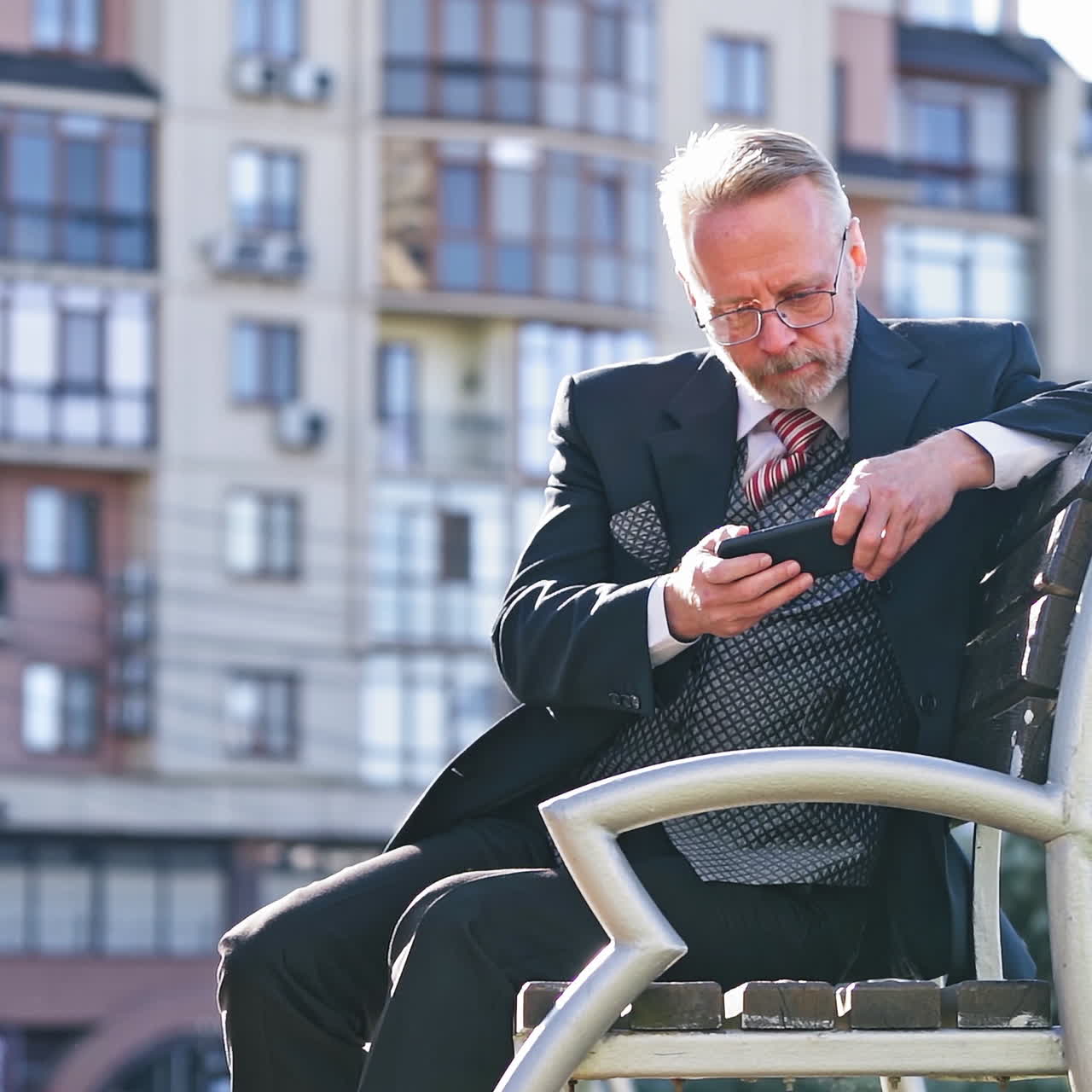 Senior businessman looking into his phone outdoors. Middle aged man wearing glasses in elegant suit sitting on a bench with a mobile phone on the high-rise building background.