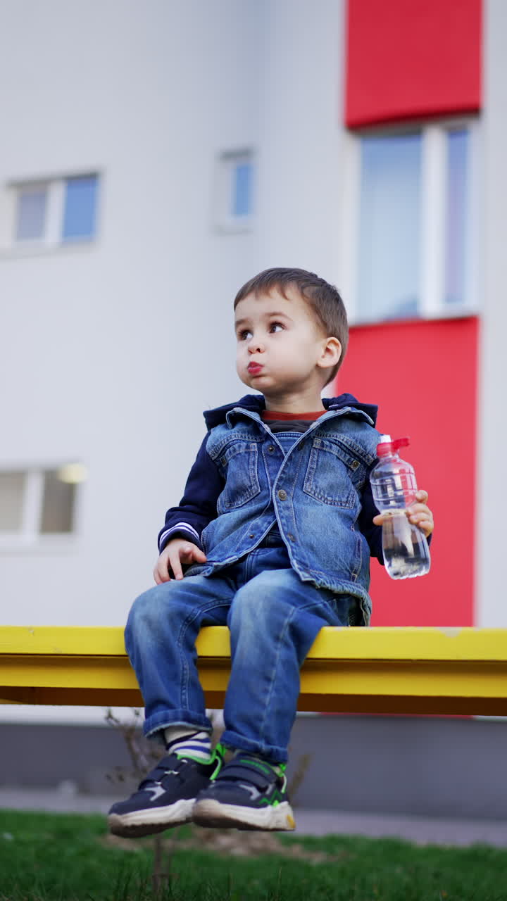 Caucasian toddler drinking water from water. Kid puts the bottle on the bench and looks around. Vertical video.