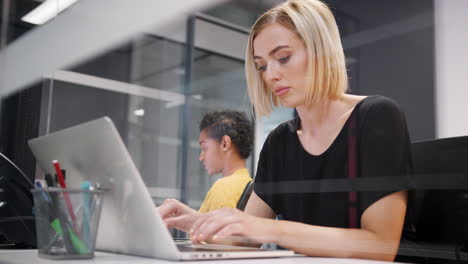 Two-young-women-working-together-on-computers-in-a-glass-office-cubicle,-one-turning-around-to-the-other,-low-angle,-close-up,-seen-through-glass