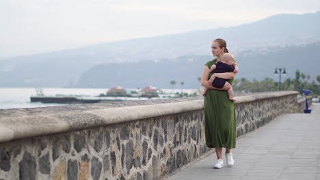 walking near the ocean on an old european square, a young mother enjoys the presence of her baby son, looking at the waves and smiling with affection