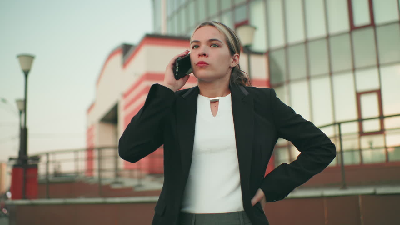 Tired white lady in professional outfit talking on phone with weary expression, walking near modern building, distant people blurred in background
