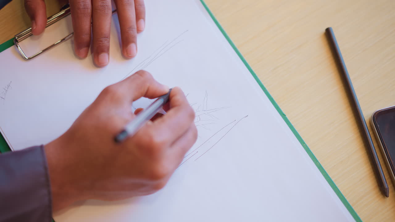 Person sketching plant on white paper using pen while other hand rests on clipboard, with pencil and phone on desk, showing creative focus, artistic expression, and planning process in calm workspace
