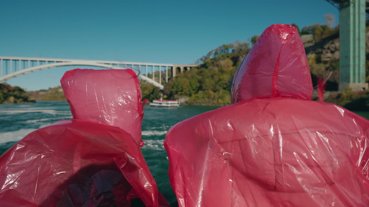 Turisti con poncho antipioggia rossi su una barca vicino alle Cascate del Niagara con il Rainbow Bridge sullo sfondo.